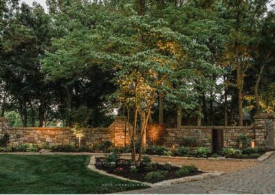 Stone wall adorned with climbing vines, surrounded by lush greenery and tall trees. Elegant stone pathway leads to a gated entrance, accentuated by warm lighting. Manicured lawn in the foreground enhances curb appeal, creating a serene garden atmosphere perfect for outdoor living spaces.