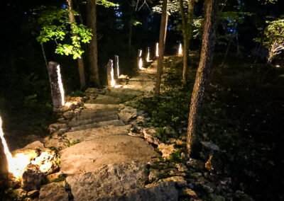 A meticulously crafted stone pathway illuminated by soft ground lights winds through a dark, wooded area, enhancing the outdoor living spaces. Tall trees and scattered vegetation create a serene, mysterious atmosphere, showcasing the elegance of landscape design in Kansas City.