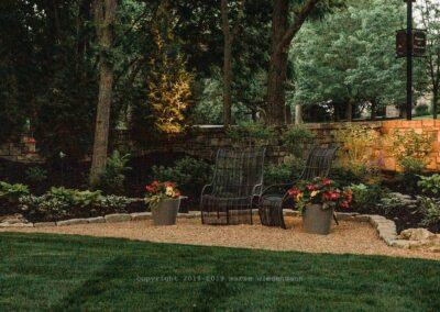 A serene garden scene showcases two black metal chairs on a gravel area, framed by lush green foliage and flowers in pots. Trees and a stone wall form the backdrop, while soft lighting enhances the tranquil atmosphere—a perfect example of outdoor living spaces.