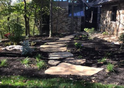 The stone pathway, enhancing the curb appeal, winds through a mulched garden with young plants and trees alongside. In the background, a modern house with stone and glass elements is partially visible. Sunlight filters through the trees, casting enchanting shadows.