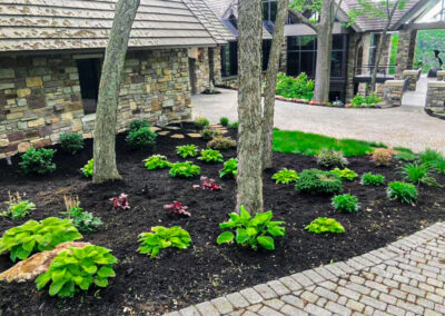 A beautifully landscaped garden with lush greenery and various plants adds stunning curb appeal to the stone house. The mulched areas around the trees and a paved stone path that curves through this charming outdoor living space create an inviting approach to the rustic building in the background.