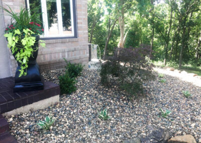 A landscaped garden with plants nestled in a bed of small rocks enhances the curb appeal. A tall black planter with green foliage and red flowers stands by a brick building. Trees and a wooded area form the background, while a small bush and several tiny plants grace the foreground.