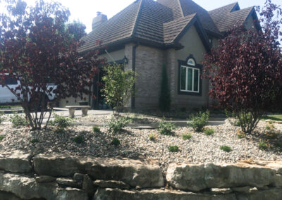 A charming house with a steep gabled roof and brick facade, surrounded by expertly crafted outdoor living spaces. In the foreground, a stone wall with decorative gravel and young trees frames the scene, showcasing sophisticated landscape design in Kansas City under a clear, sunny sky.