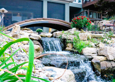 A small stone waterfall surrounded by rocks and greenery graces this serene outdoor living space. A wooden bridge arches above it, beside a potted plant with red flowers on the right. In the background, a building with large windows opens to a charming patio area, enhancing curb appeal.