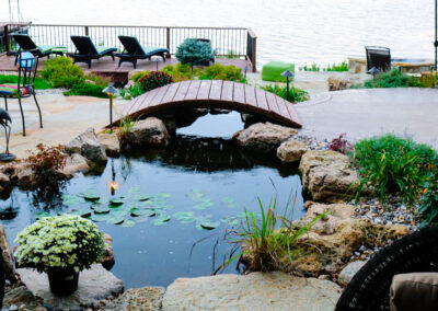 A tranquil garden, perfect for outdoor living spaces, features a small pond with lily pads, surrounded by rocks and plants. A wooden bridge arches over the pond. In the background, lounge chairs face a large body of water, enhancing curb appeal in this serene setting.