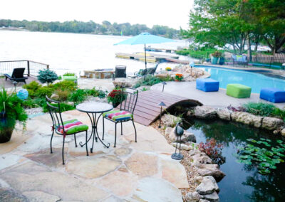 Patio with colorful cushioned chairs and a small table, overlooking a serene lake. A wooden bridge crosses a pond with lily pads. Embracing outdoor living spaces, nearby, there are blue cube seats, a swimming pool, and a large umbrella. Trees and lush greenery enhance the landscape design in Kansas City.