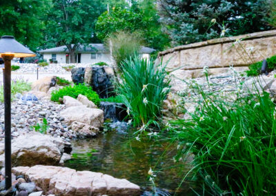 A serene garden scene with a small pond surrounded by rocks and lush greenery exemplifies landscape design in Kansas City. A gentle waterfall flows into the pond, while a stone wall and house add curb appeal in the background. A warm garden lamp stands gracefully on the left side.