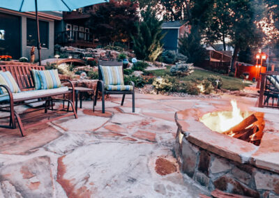 Outdoor patio with stone flooring features a fire pit surrounded by wooden chairs with striped cushions. An umbrella provides shade while garden care elevates the lush greenery and decorative lights enhance the cozy atmosphere, boosting curb appeal in this landscaped garden.