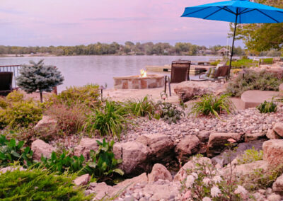 A tranquil lakeside scene at sunset features a stone fire pit surrounded by chairs, perfect for enhancing your homes curb appeal. An umbrella provides shade amidst lush greenery and rocks. A dock extends into the calm water, framed by trees in the distance.