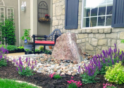 A landscaped garden with a large rock centerpiece surrounded by purple and pink flowers. Theres a black bench with red cushions beside a stone house, featuring a large window with shutters. The garden has mulched and stone areas, bordered by green grass.