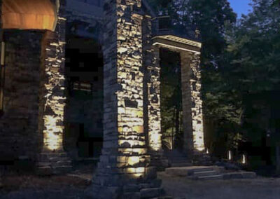 A stone building with illuminated columns at night. The structure is surrounded by trees, and the lighting creates dramatic shadows on the stonework. The sky is dark blue, indicating early evening or night.