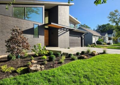 Modern house with dark brick exterior, large windows, and a slanted roof. The landscaped front yard features trimmed bushes, rocks, and a small tree. A driveway and neighboring houses are visible in the background under a clear blue sky.