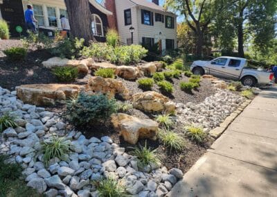 A landscaped front yard with large rocks and small shrubs on a sloped garden bed. A pickup truck is parked on a driveway beside the garden. Two people are visible near the house, which has white and brick siding. Trees and grass surround the area.