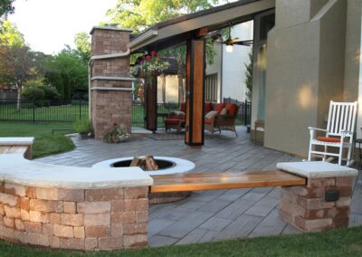 Outdoor patio featuring a fire feature with semi-circular stone seating and a wooden bench. A covered seating area is visible in the background with red cushioned chairs and hanging plants. Trees and lawn surround the space, enclosed by a black fence.