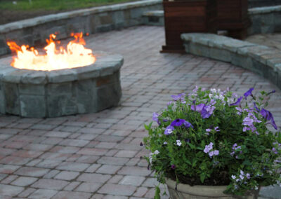 A patio with a stone fire pit on the left, flames visible. In the foreground on the right is a planter filled with purple and white flowers. The ground is paved with stone tiles. Curved stone benches are in the background.