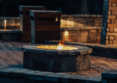 A stone fire pit surrounded by a brick patio with two wooden benches nearby. The fire emits a warm glow, and a stone wall is visible in the background. The scene suggests a cozy outdoor setting.