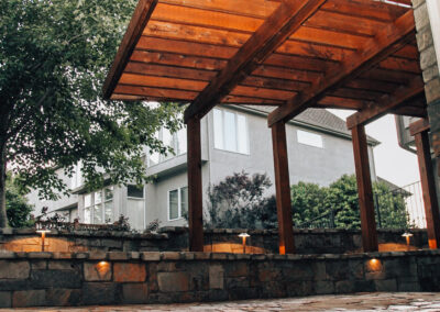 A patio with a wooden pergola and stone walls, surrounded by trees. Small lights are embedded in the walls, and a house is visible in the background under a cloudy sky.