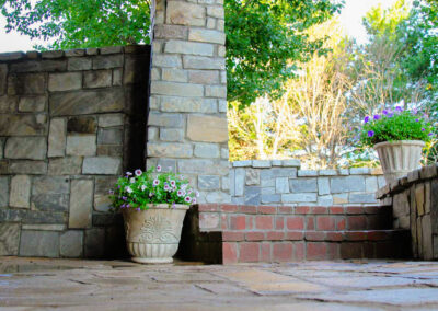 Stone patio with two planters filled with purple and white flowers. The patio features a stone wall, a brick step, and lush green trees partially visible behind the structure. Sunlight casts a warm glow across the scene.