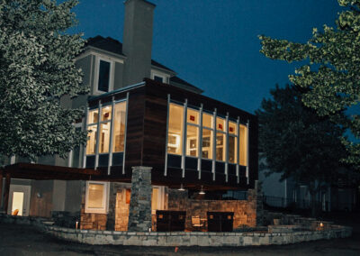 A modern house at dusk with a large, illuminated glass extension on the second floor. The structure features stone pillars and dark wood accents, surrounded by trees. The sky is a deep blue, enhancing the warm glow from the windows.