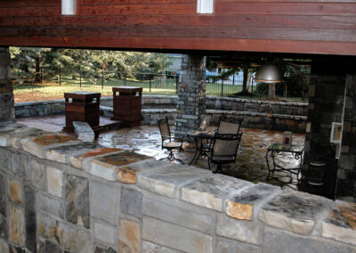 Outdoor stone patio with a rustic design featuring a round metal table and chairs. There are stone pillars, wooden accents, and a large metallic lamp. The area is surrounded by greenery and a fence.