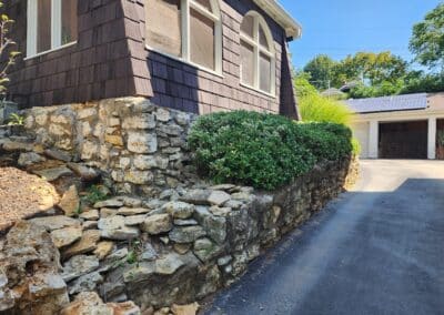 A house with brown shingle siding sits on a small hill, supported by a stone retaining wall. Bushes are planted along the wall. A paved driveway leads to a garage with solar panels on its roof. Trees and a clear blue sky are in the background.